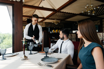 A young sommelier in a stylish apron demonstrates and offers fine wine to a beautiful couple in a restaurant. Customer service in an elite restaurant and a public restaurant.