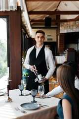 A young sommelier in a stylish apron demonstrates and offers fine wine to a beautiful couple in a...