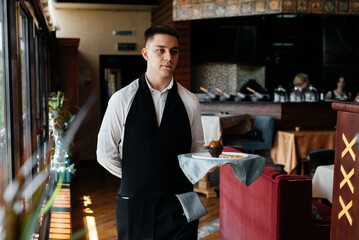 A young waiter in a stylish uniform stands with an exquisite dish on a tray near the table in a beautiful restaurant close-up. Restaurant activity, of the highest level.