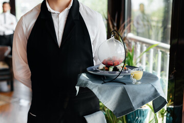 A young waiter in a stylish uniform stands with an exquisite dish on a tray near the table in a...