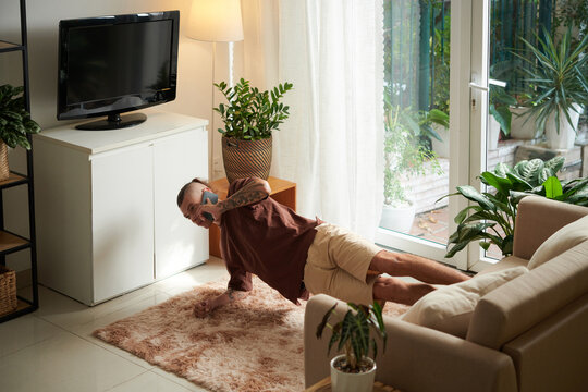 Fit young man standing in side plank with legs on sofa and talking on phone