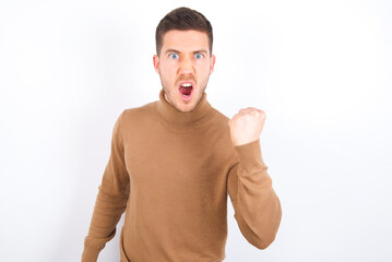 young caucasian man wearing grey turtleneck over white background angry and mad raising fist frustrated and furious while shouting with anger. Rage and aggressive concept.