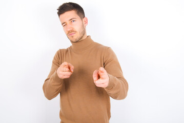 Joyful young caucasian man wearing grey turtleneck over white background wink and points index...
