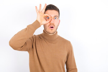 young caucasian man wearing grey turtleneck over white background doing ok gesture shocked with surprised face, eye looking through fingers. Unbelieving expression.