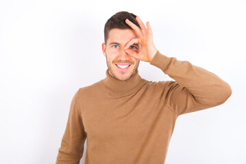 young caucasian man wearing grey turtleneck over white background doing ok gesture with hand smiling, eye looking through fingers with happy face.