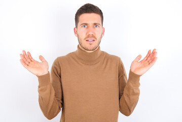 Fototapeta premium Indignant young caucasian man wearing grey turtleneck over white background gestures in bewilderment, frowns face with dissatisfaction.