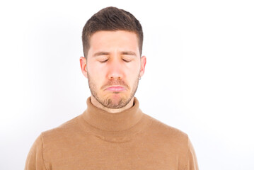 Obraz premium Dismal gloomy rejected young caucasian man wearing grey turtleneck over white background has problems and difficulties, curves lower lip and closes eyes in despair, being in depression