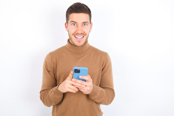 Excited young caucasian man wearing grey turtleneck over white background holding smartphone and looking amazed to the camera after receiving good news.