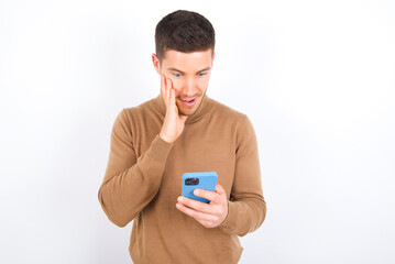 young caucasian man wearing grey turtleneck over white background holding in hands cell reading sms using new app 5g