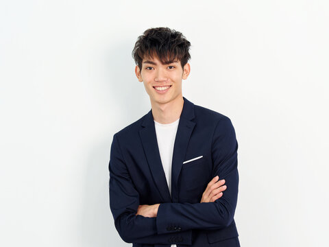 Portrait Of Handsome Chinese Young Man In Dark Blue Leisure Suit Posing Against White Wall Background. Arms Crossed With Mobile Phone In Hand And Looking At Camera, Looks Confident, Front View