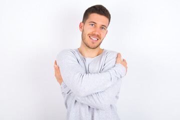 Charming pleased young caucasian man wearing grey sweater over white background embraces own body, pleasantly feels comfortable poses. Tenderness and self esteem concept