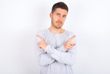 young caucasian man wearing grey sweater over white background crosses arms and points at different sides hesitates between two items or variants. Needs help with decision