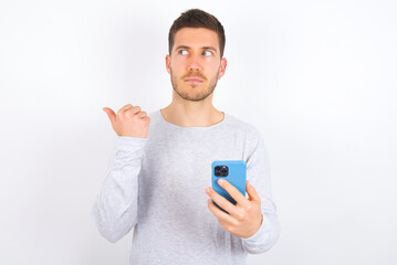 young caucasian man wearing grey sweater over white background points thumb away and shows blank space aside, holds mobile phone for sending text messages.