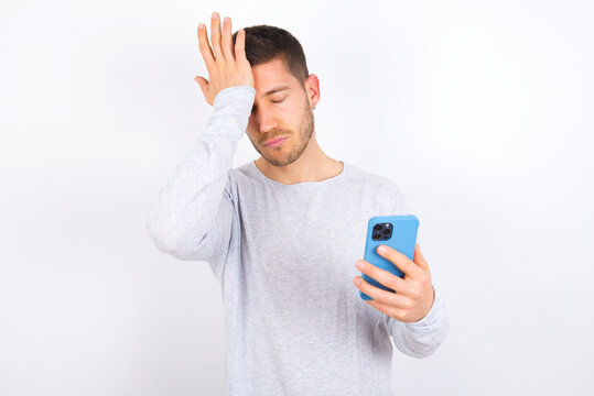 Upset depressed young caucasian man wearing grey sweater over white background makes face palm as forgot about something important holds mobile phone expresses sorrow and regret blames