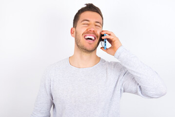Overemotive happy young caucasian man wearing grey sweater over white background laughs out positively hears funny story from friend during telephone conversation