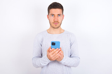 Portrait of serious confident young caucasian man wearing grey sweater over white background holding phone in two hands