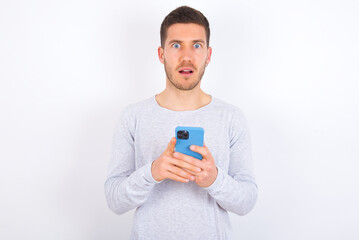 Photo of astonished crazy young caucasian man wearing grey sweater over white background hold smartphone dislike feedback concept