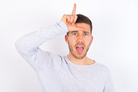 Funny young caucasian man wearing grey sweater over white background makes loser gesture mocking at someone sticks out tongue making grimace face.