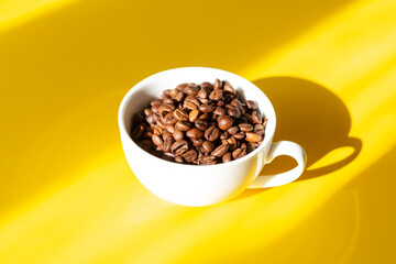 Coffee beans in a white ceramic cup on a yellow background. Hard sun shadows