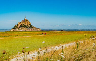 Tourists crossing empty green fields to visit the famous Le Mont Saint Michel tidal island on a...