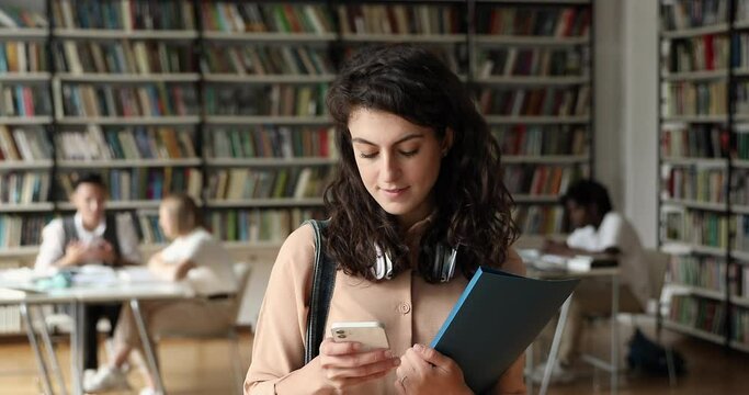 Young Latin woman student stand in college library apart of groupmates using mobile phone search information online. Happy smiling teen lady take break in learning read message in chat on cell screen