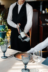 A young sommelier in a stylish uniform demonstrates and offers the client fine wine in the restaurant. Customer service. Table setting in a fine restaurant.
