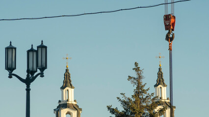 Red hook of a crane on the background of the tower church with orthodox cross. Construction site.