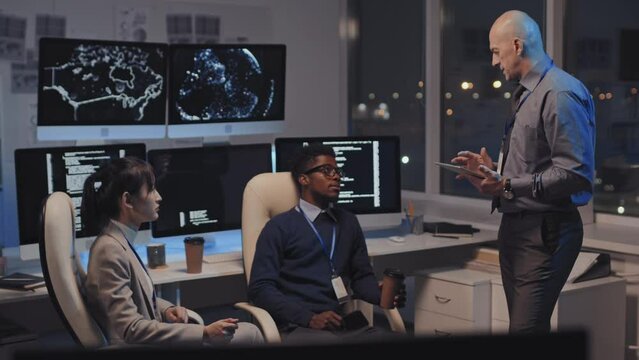 Medium long of Caucasian male IT department manager using tablet, giving tasks to two diverse employees, sitting at desk with monitors in office at night, Asian woman and Black man nodding