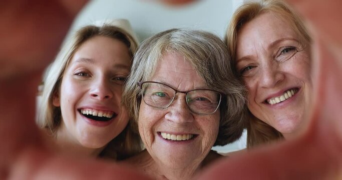Diverse cheerful Multigenerational women close up front view of smiling faces looking at camera through joined fingers showing heart shape hand sign gesture. Concept of family ties, bonding and love