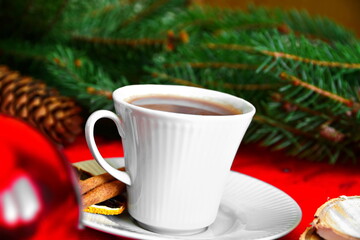 Obraz na płótnie Canvas Winter drink. Cup of hot chocolate or coffee or cocoa close up decorated with cinnamon sticks and slice of dry lemon on white soucer, twig of christmas tree, cones and red ball on blurred background