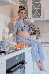 A pregnant girl in pajamas in a good mood drinks tea with cookies in the kitchen.