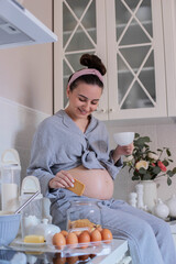A pregnant girl in pajamas in a good mood drinks tea with cookies in the kitchen.