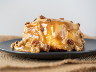 Close-up of a cinnamon bun topped with caramel topping and pecans. Black plate, side view. Delicious sweet dessert, blurred background. Selective focus