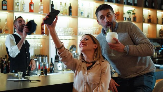 Cheerful couple with glasses of cocktails sitting in bar and making selfie on phone