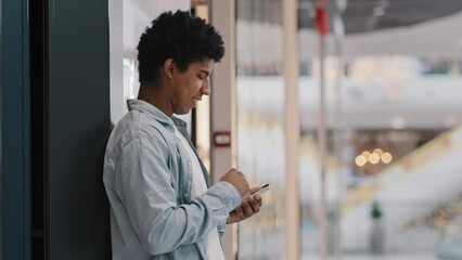 African american millennial man biracial guy stands near wall indoors swipes into phone screen mobile chat app chooses online shopping with smartphone reads news browsing telephone free wifi side view - Powered by Adobe