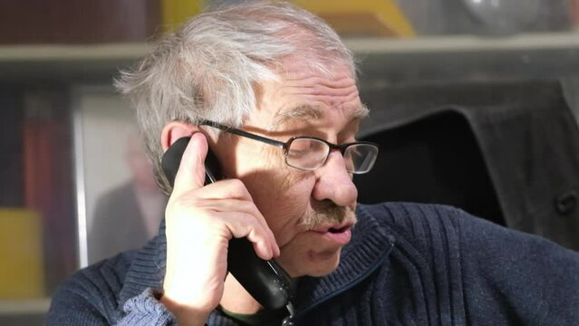 An elderly man in glasses with a mustache makes excuses and is indignant while talking on a landline phone. Elderly engineer calling on the phone