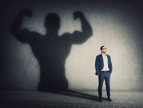 Confident businessman keeps hands in his pockets while casts a powerful person shadow on the wall behind. Business person transformation as metaphor for leadership and motivation