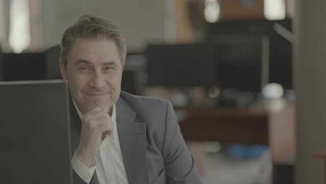 Business portrait - confident businessman sitting at desk in office. Happy mid adult man in shirt and jacket, smiling. Bearded, gray hair. Video in LOG format - S-LOG3 Cine.