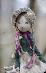 A charming beige doll kitty with green eyes dressed in a jacket, dress and cap sitting on a tree and looking sideways.