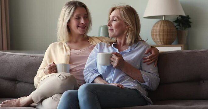 Young woman her older mom sit on sofa and talking, having pleasant conversation, enjoy beverage in cups take coffee break share news. Family bond, trust between mother and grown up daughter concept