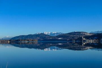 View on Maria Worth and Woerthersee from Poertschach in Carinthia, Austria. Calm lake reflecting the landscape. View on Koschutnikturm and Hochobir. Village with church at the waters edge. Sunny. Alps