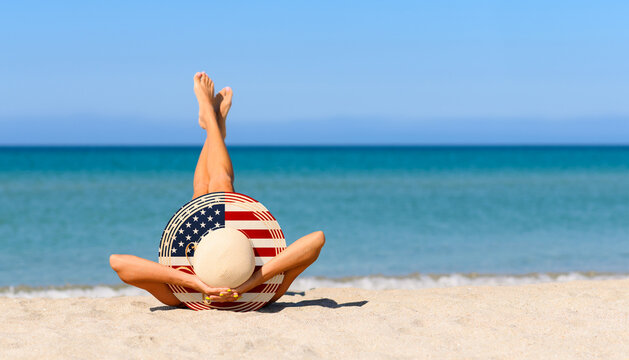 A slender girl on the beach in a straw hat in the colors of the USA flag. The concept of a perfect vacation in a resort in the United States. Focus on the hat. - Powered by Adobe
