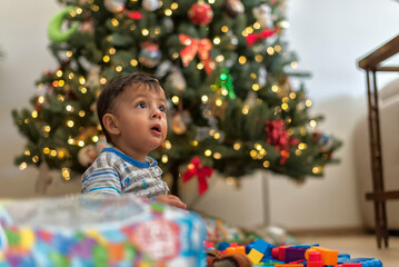 Fototapeta na wymiar Little latin toddler playing with his christmas presents by the tree