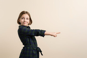 Joyful Little Girl 9s in Plaid Shirt Dress Points Index Finger in ivory Background. Smart Child Have Excellent Idea. Showing promo. Inviting use link. Give direction.