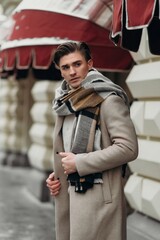  Young man wearing coat walking in the street. Young bearded guy with modern hairstyle in urban background
