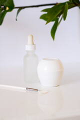 White jar of cream and bottle with a dropper from serum with a brush for applying a face mask. Green leaves in a frame. Simple organic eco friendly moisturizing products. Skin disease, aging, Wilting.