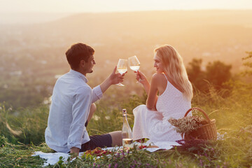 Loving young couple enjoying picnic at sunset and is clinking wine glasses during romantic date.
