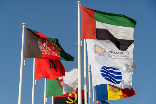 Many world flags flying in a row outside of expo center in 2020 waving on blue sky background with UAE, Afganistan, Expo 2020 and other flags.