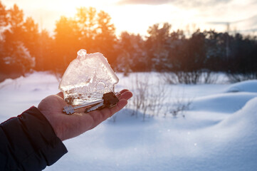 man's hand holding small glass, crystal house and keys in  palm of his hand against background of winter landscape. Artistically colored and tinted photo. Concept dream home, take mortgage. Copy space