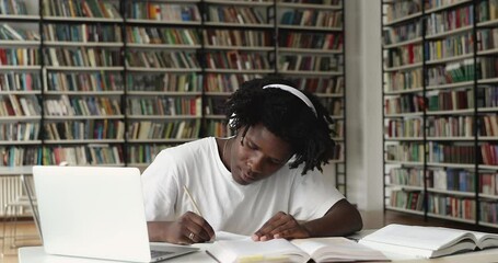 Diligent young Afro American male in earphones study paper literature at contemporary library. Smart guy student teenager prepare for examination test write essay summary assignment search information